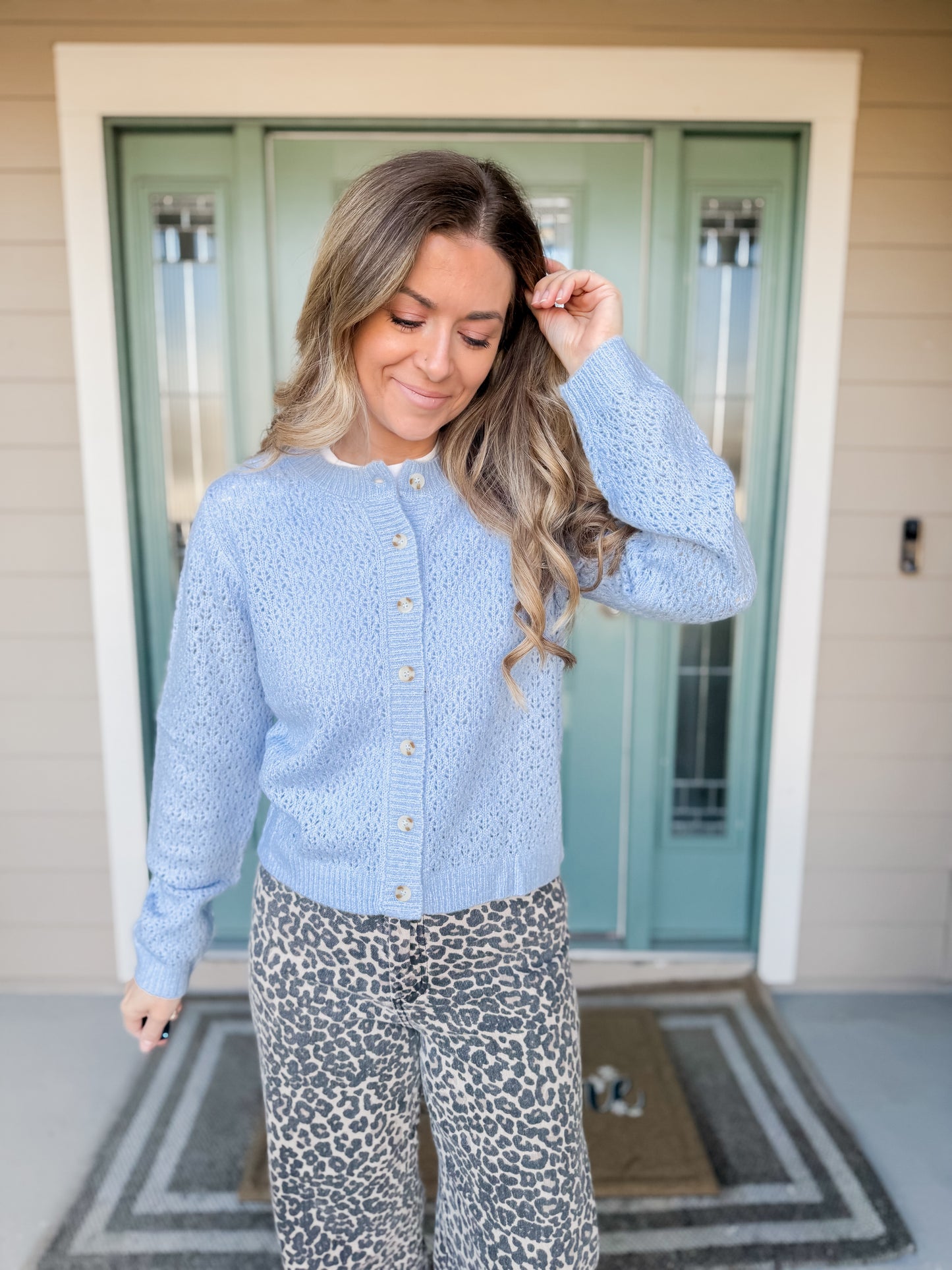 Woman wearing a light blue cardigan and leopard print pants standing in front of a green door. haven eyelet knit cardigan.