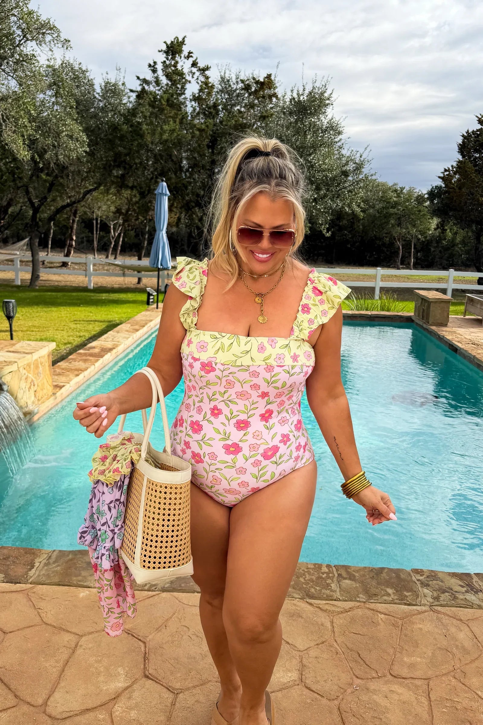 Woman in a floral swimsuit standing by a pool with trees and a fence in the background