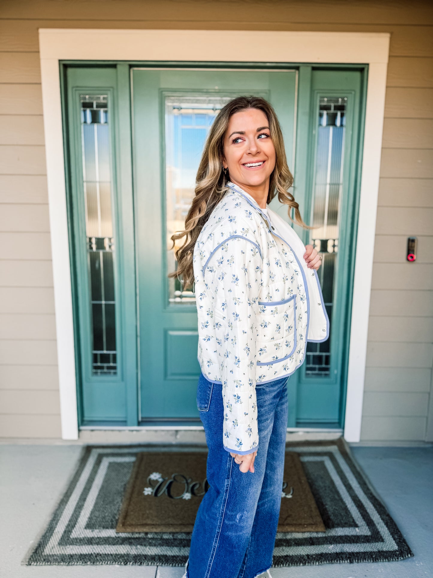 Woman standing in front of a teal door wearing a white floral jacket with blue trim and blue jeans, side view. longsleeve 