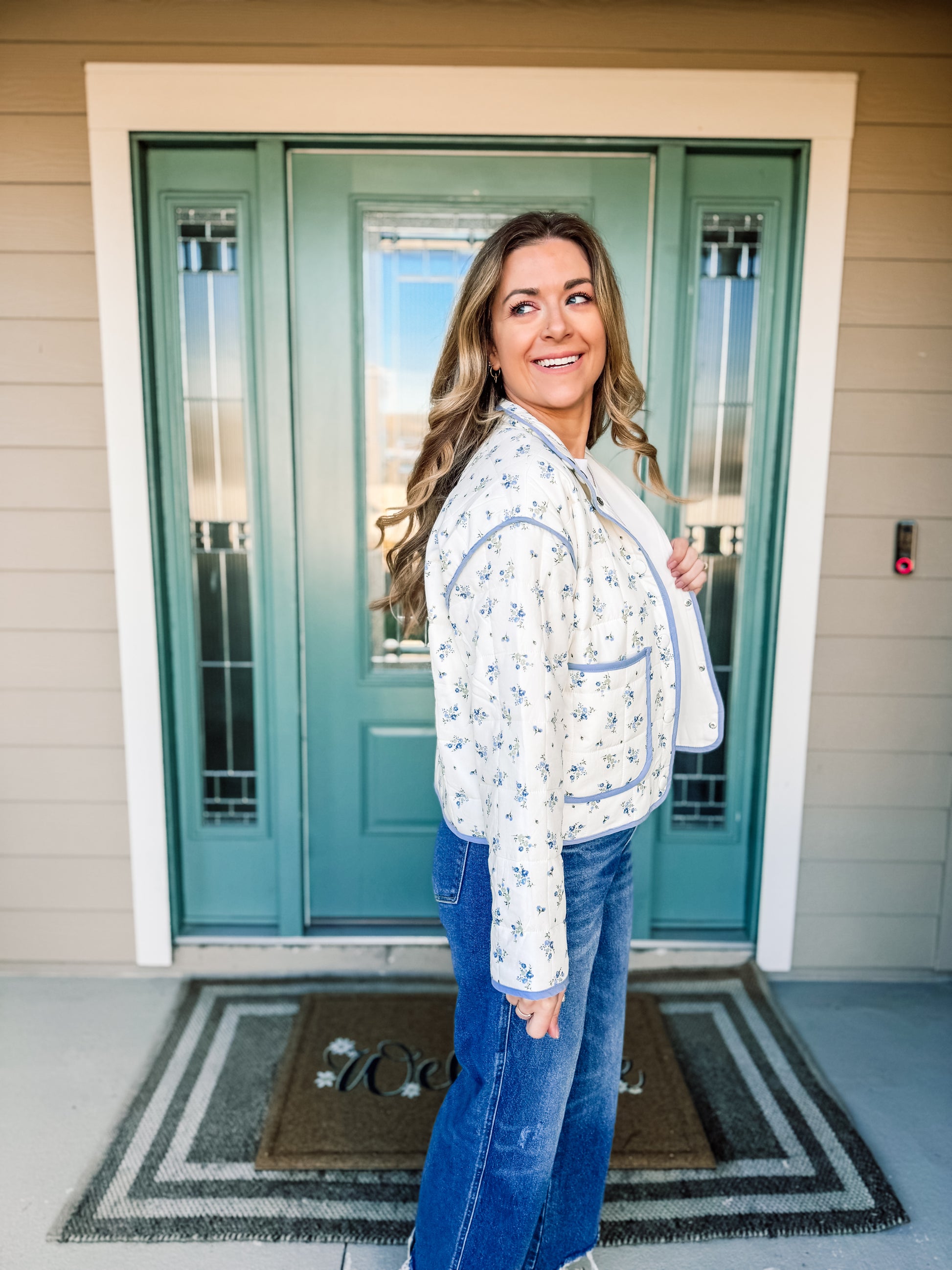 Woman standing in front of a teal door wearing a white floral jacket with blue trim and blue jeans, side view. longsleeve 