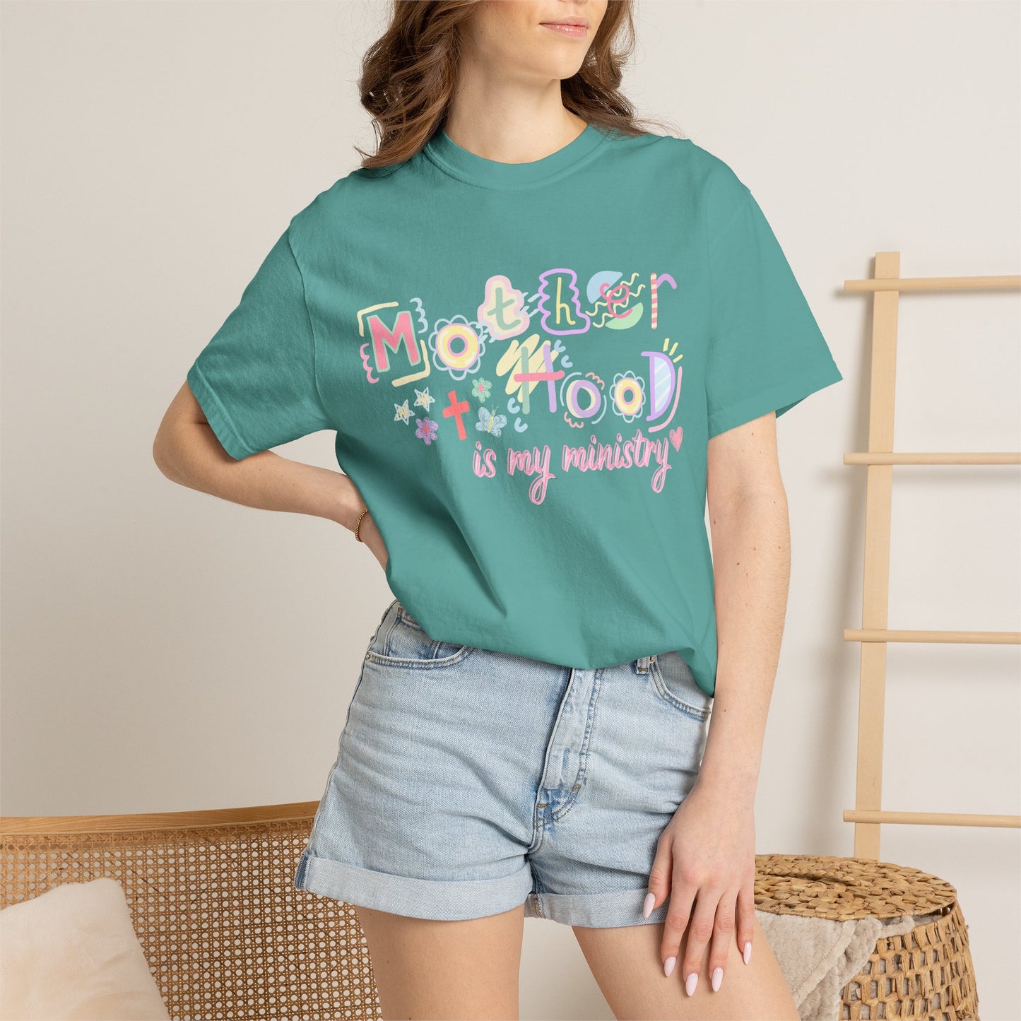 Person wearing a teal t-shirt with colorful text "motherhood is my ministry" design in a casual setting