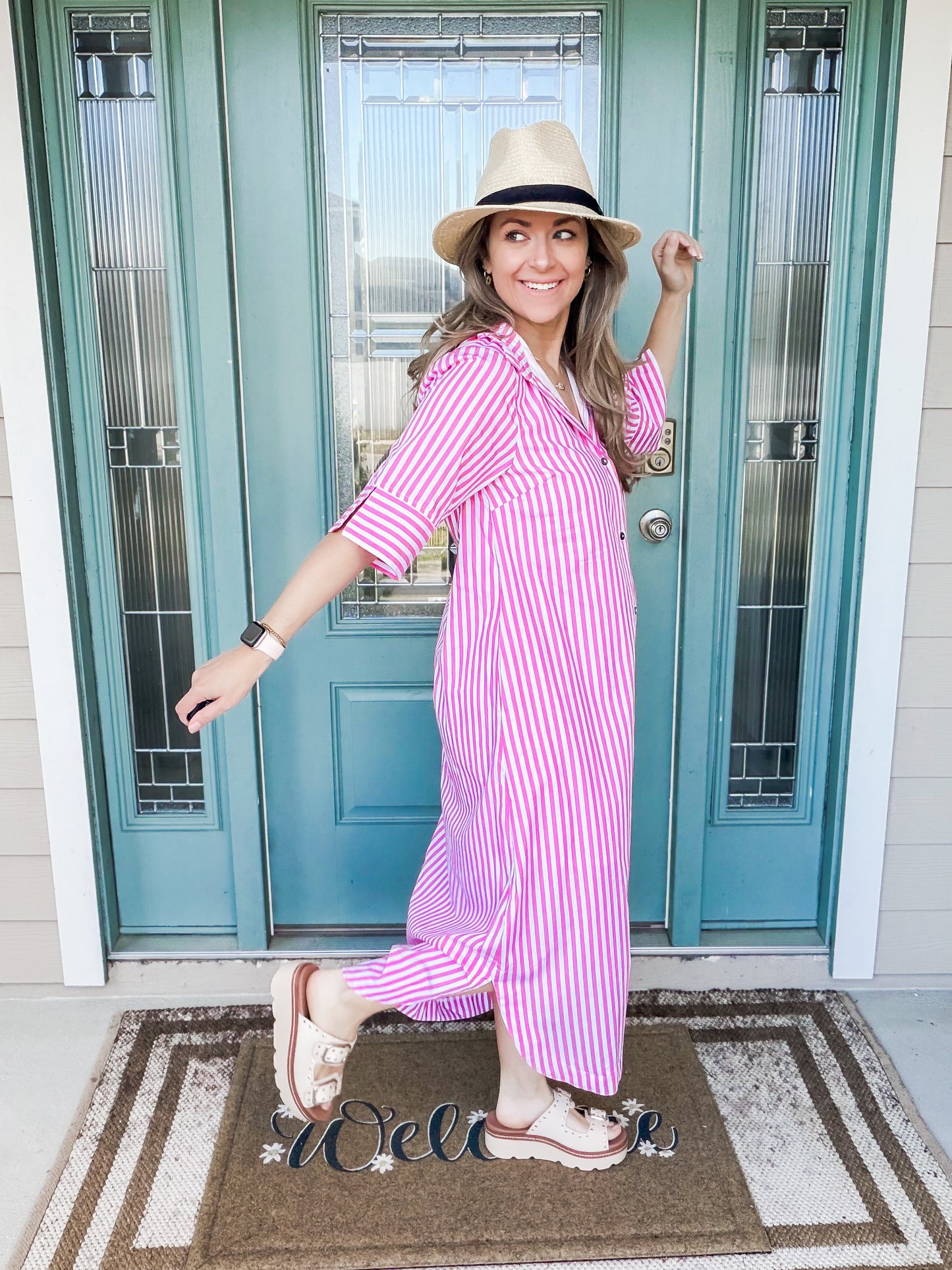 Woman in a pink and white striped dress and hat standing in front of a teal door.