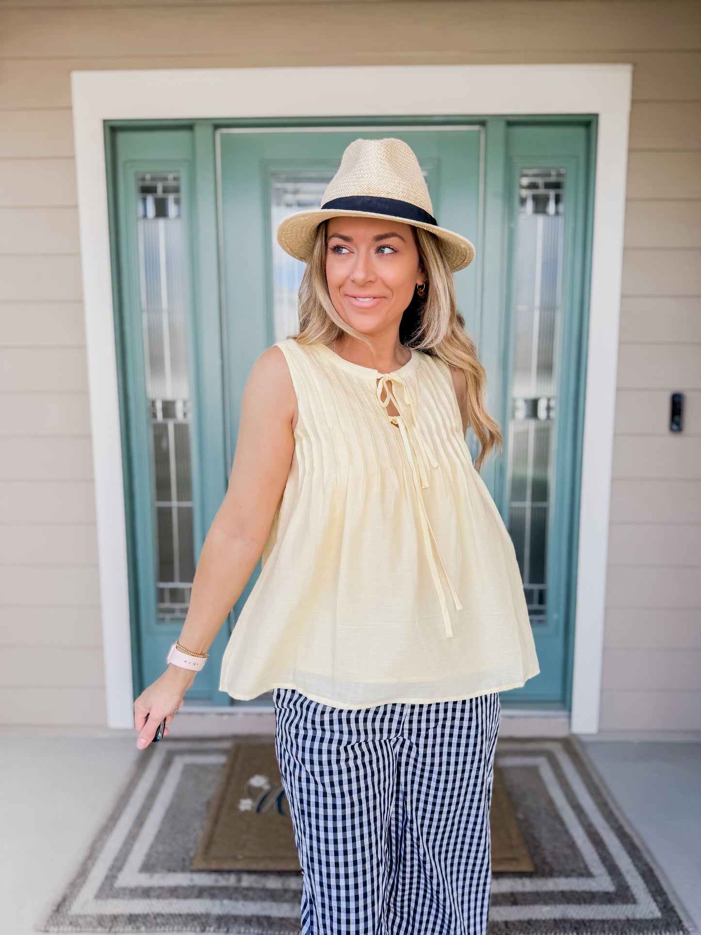 Woman wearing a yellow short sleeve pintuck top showing airy feel and checkered pants on a porch.
