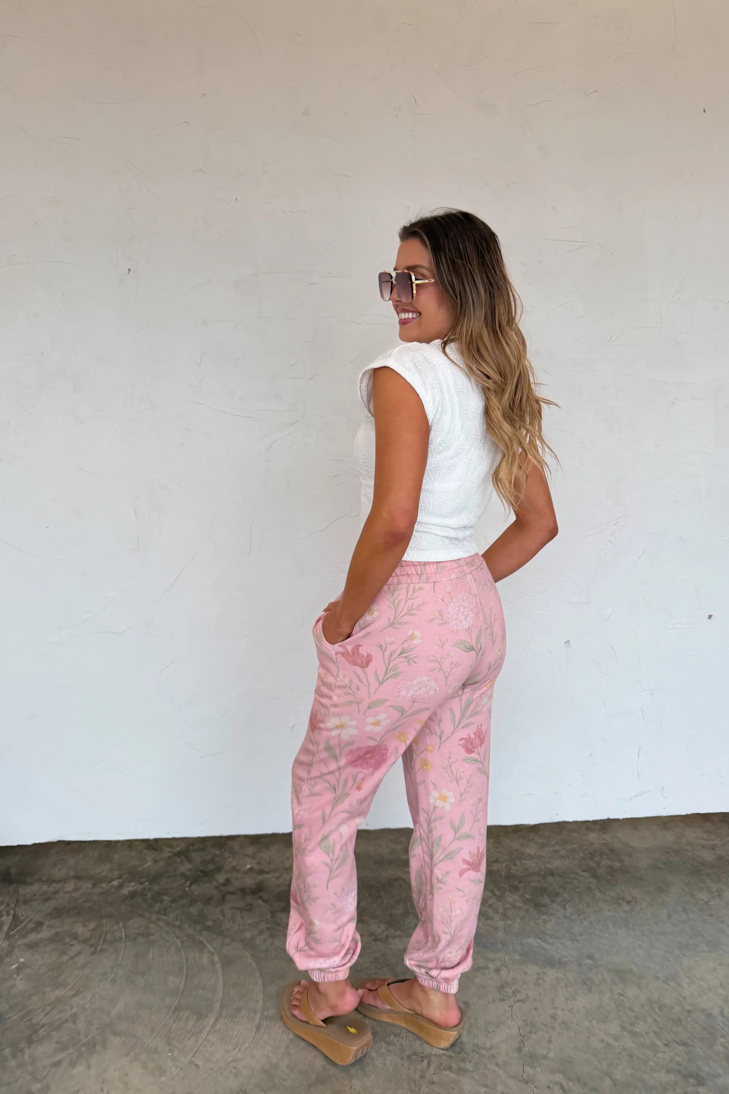 Woman wearing pink floral pants and a white top against a plain wall.