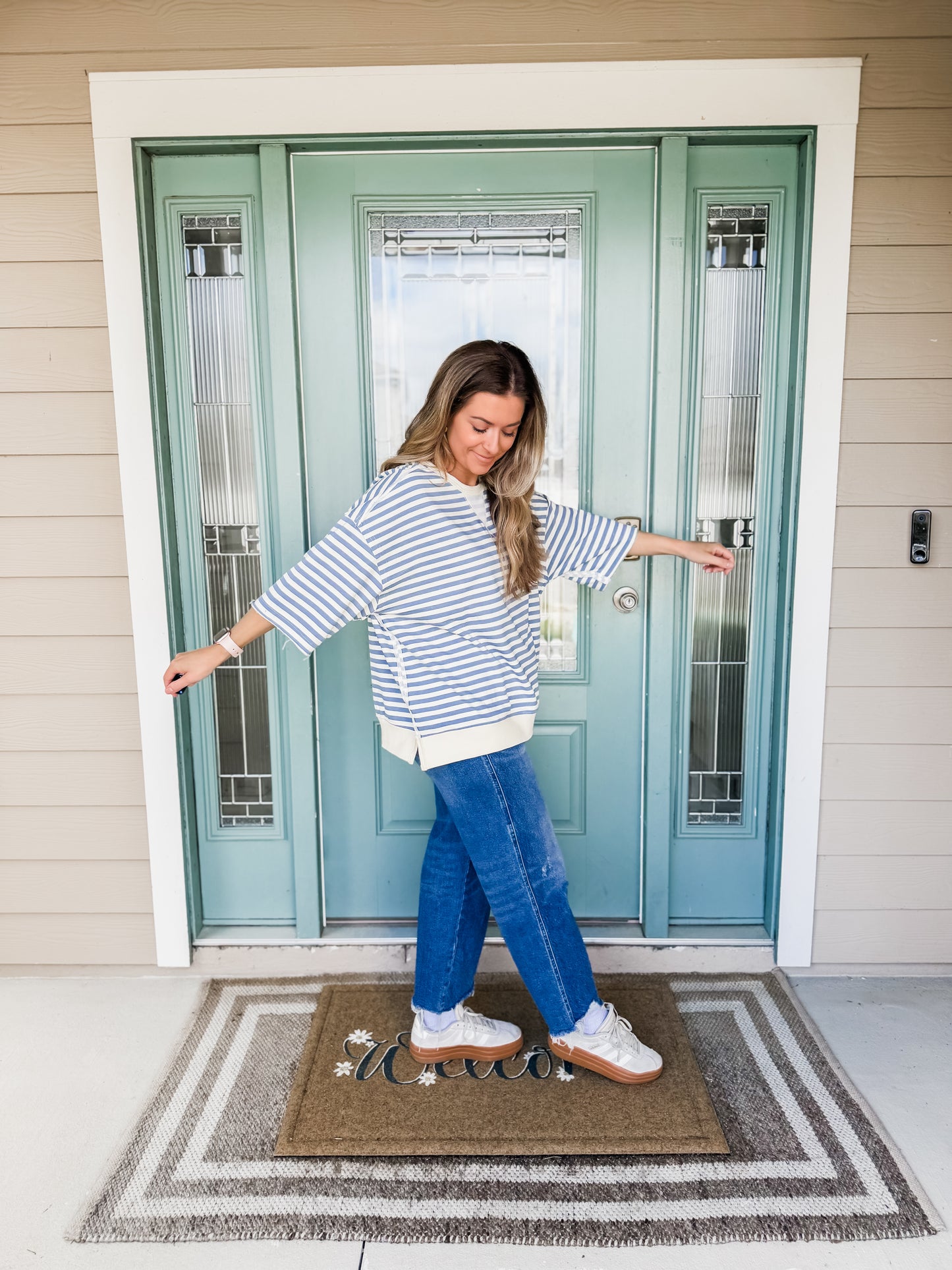 Woman standing on a doormat in front of a teal door, wearing a blue and white striped oversized french terry shirt & jeans. 