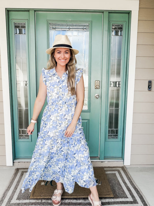 Woman in a blue long floral dress and hat standing in front of a teal door.