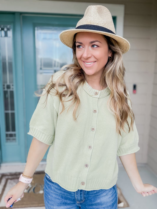 Woman wearing a light green knit sweater top with buttons and blue jeans with a straw hat.