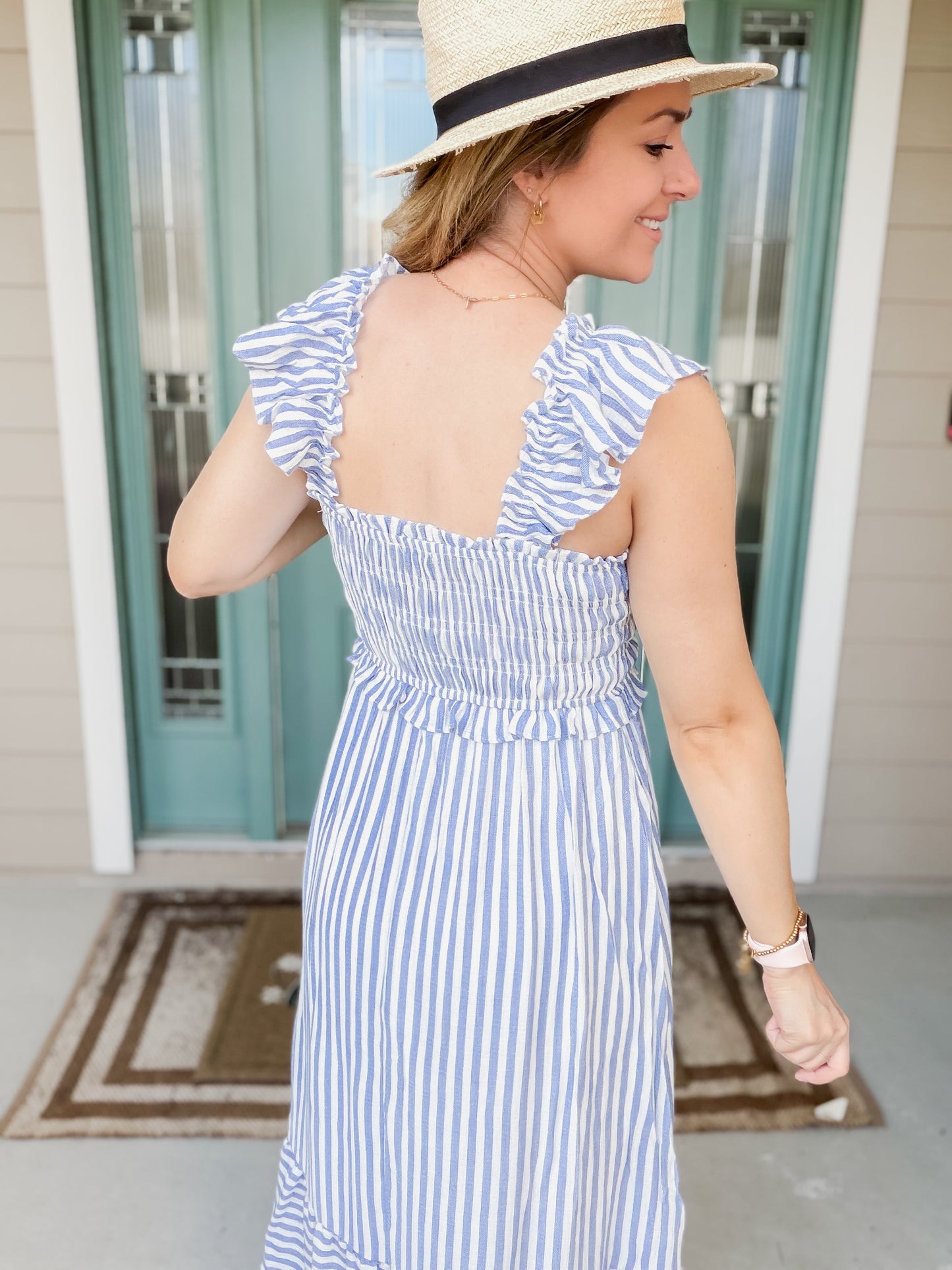 Woman wearing a blue and white striped dress with a straw hat on a porch backview shows ruffle strap detail. 
