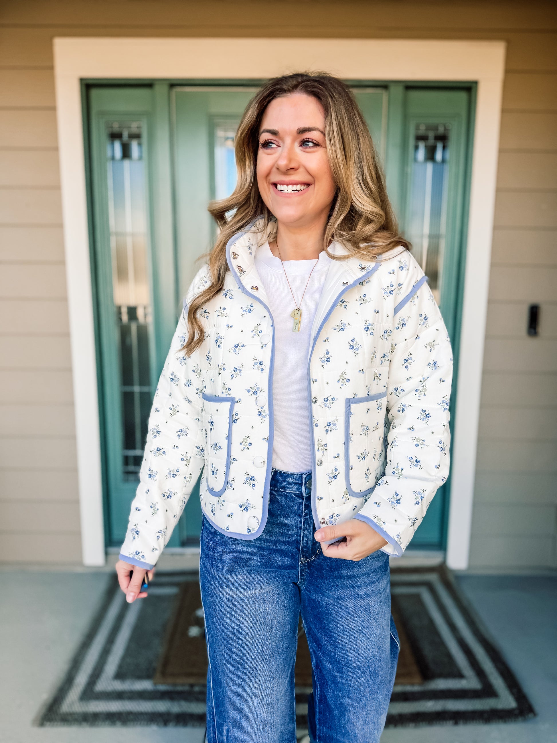 Woman wearing a floral jacket with blue trim and jeans standing in front of a door. Bloom in Blue Quilted Jacket