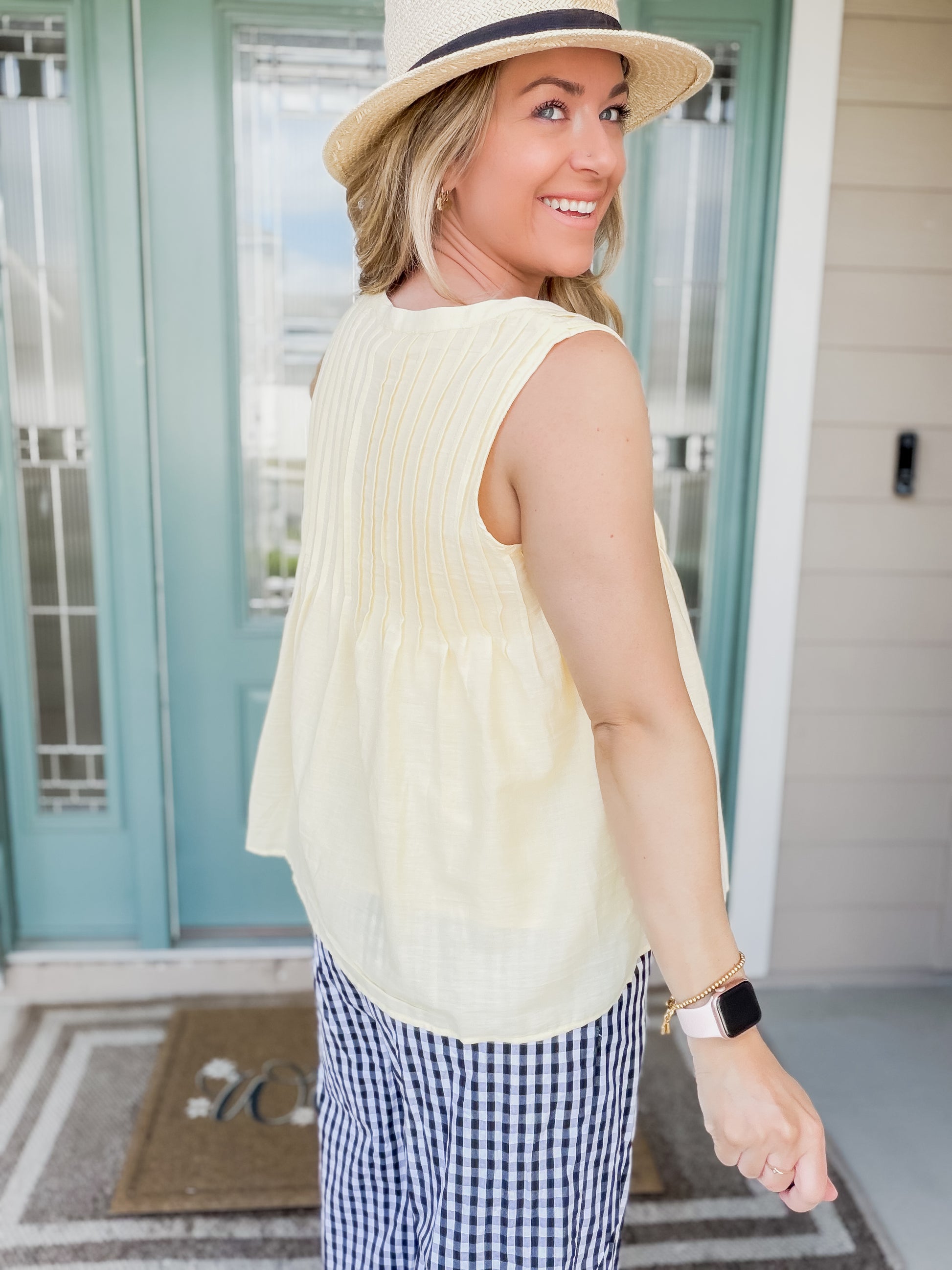 Woman wearing a yellow short sleeve pintuck top back view and checkered pants on a porch.