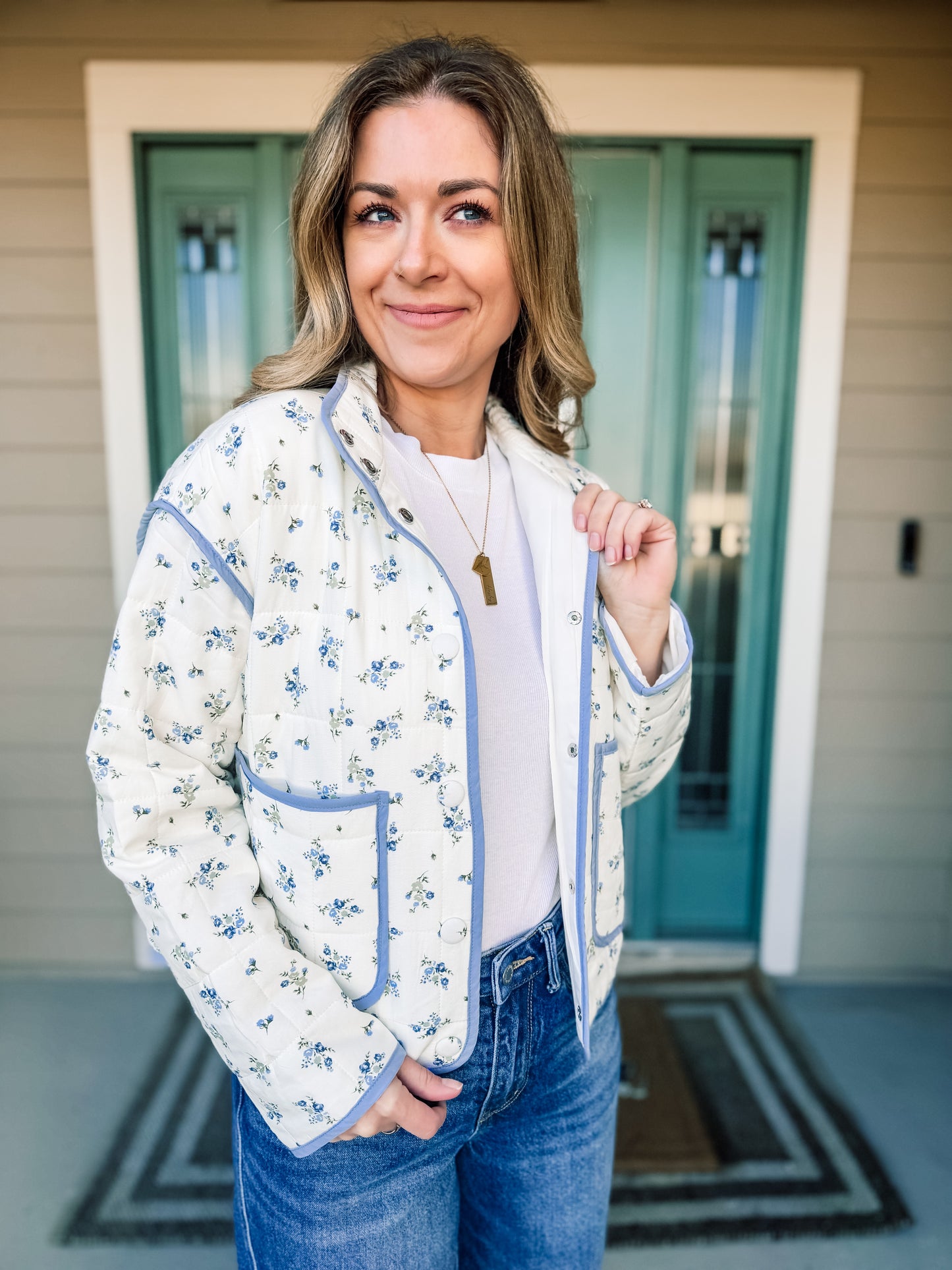 Woman wearing a floral quilted jacket  with blue trim, in front of a house with a teal door. product named Bloom in Blue Quilted Jacket