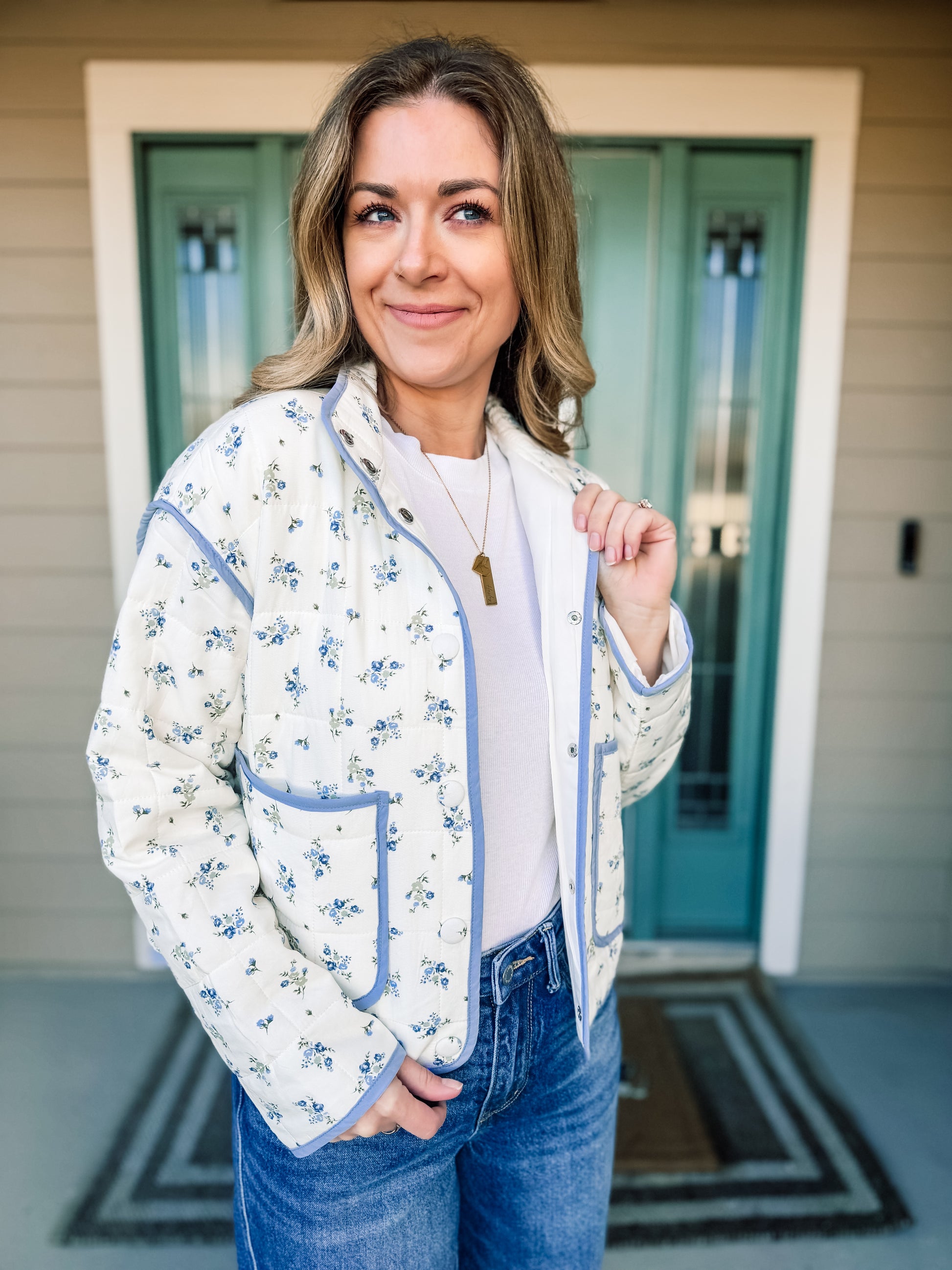 Woman wearing a floral quilted jacket  with blue trim, in front of a house with a teal door. product named Bloom in Blue Quilted Jacket
