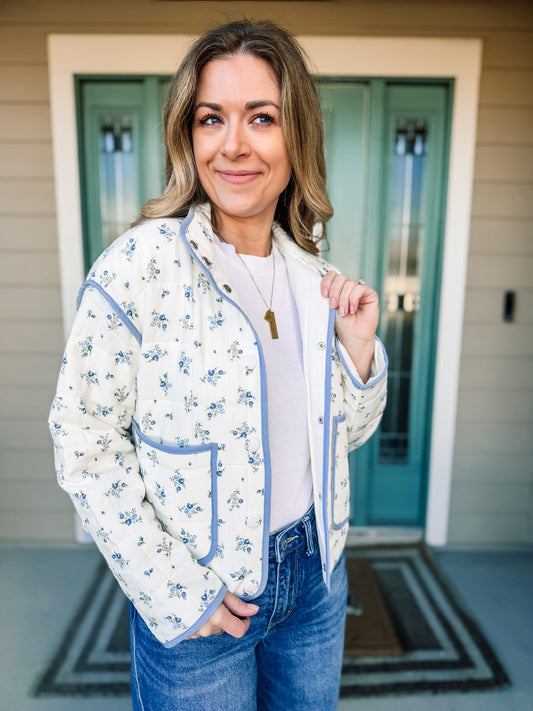 Woman wearing a floral quilted jacket  with blue trim, in front of a house with a teal door. product named Bloom in Blue Quilted Jacket