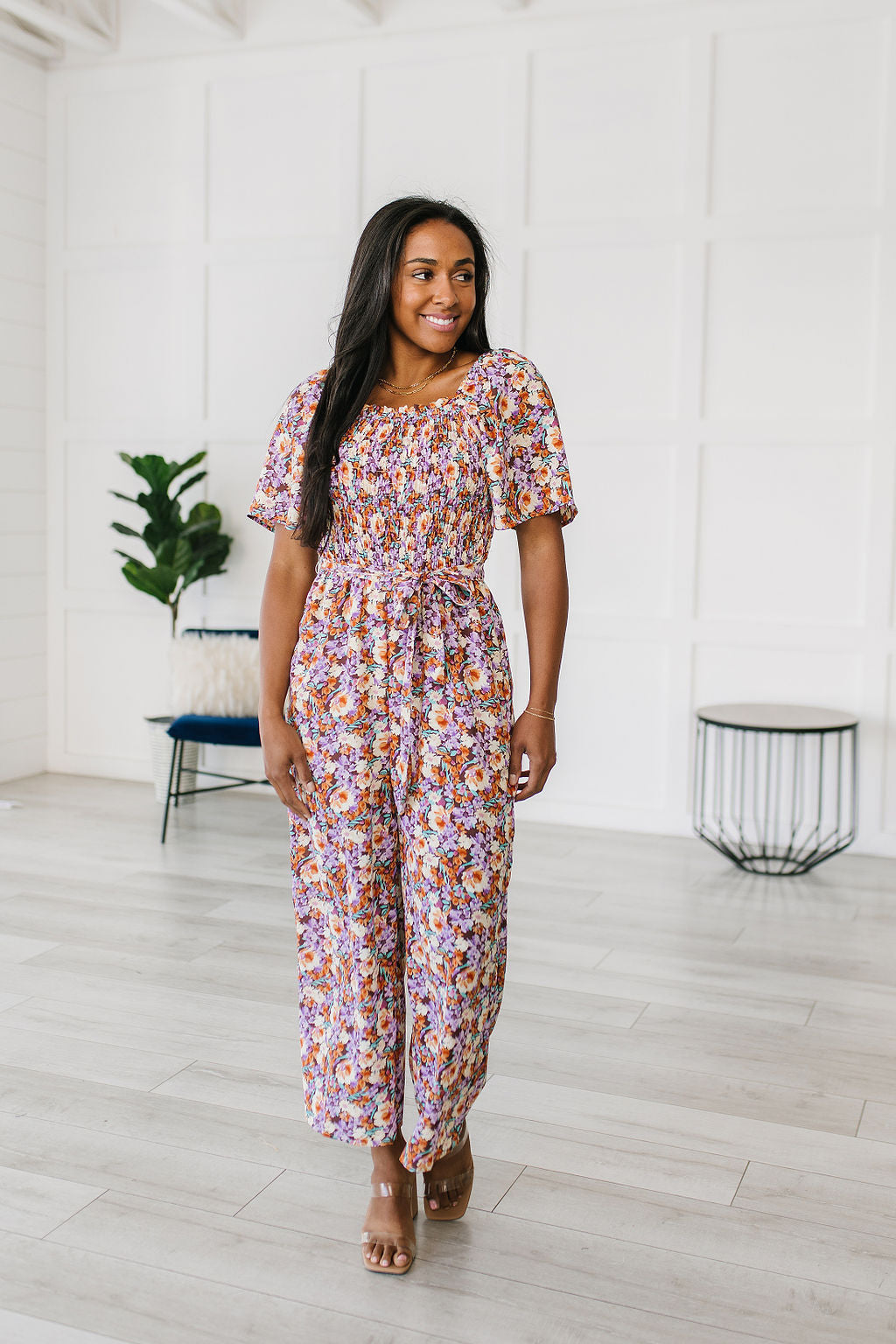 Woman wearing a floral jumpsuit standing in a minimalistic room with white walls and light wood flooring.