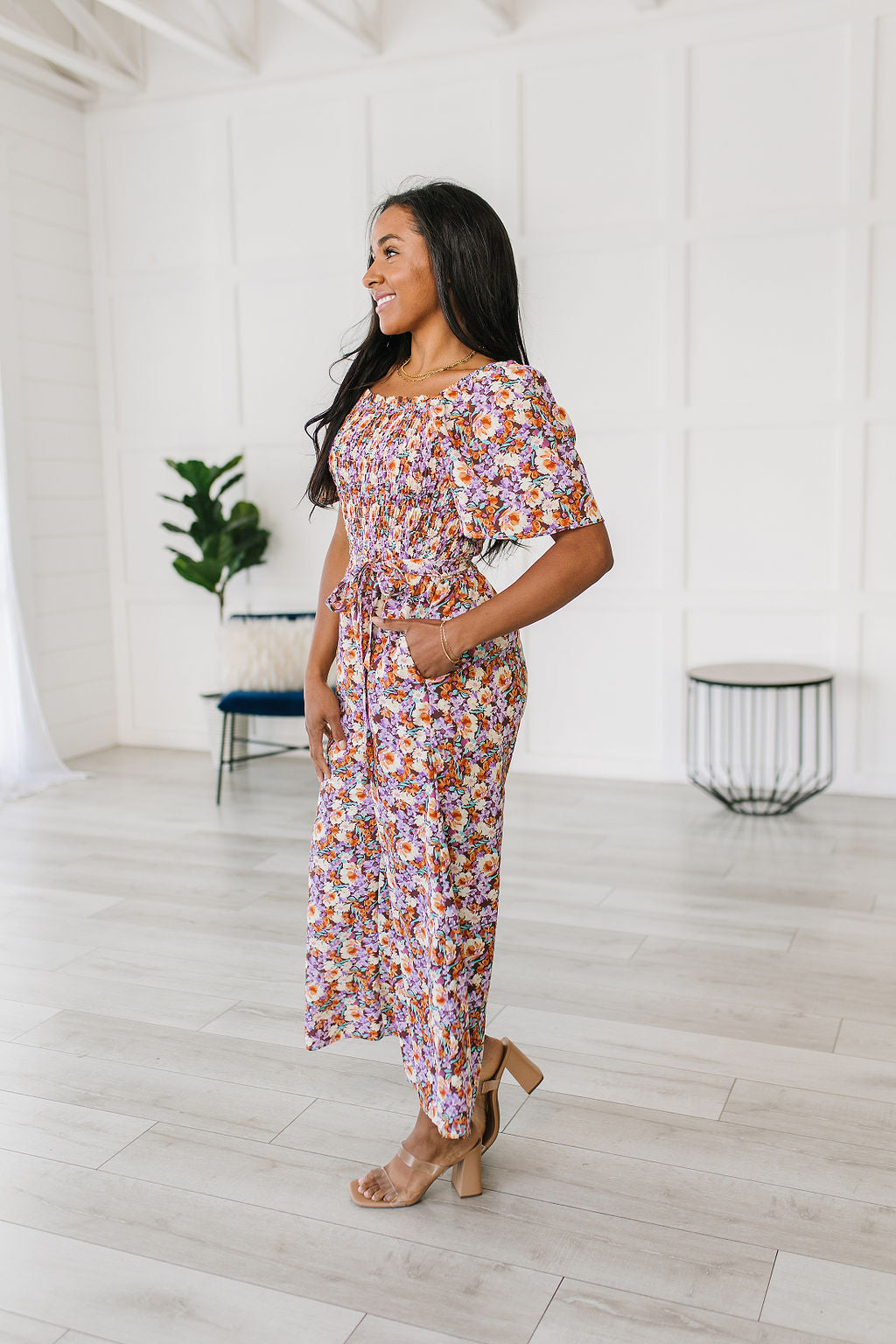 Woman wearing a floral jumpsuit standing in a minimalistic room with white walls and light wood flooring.