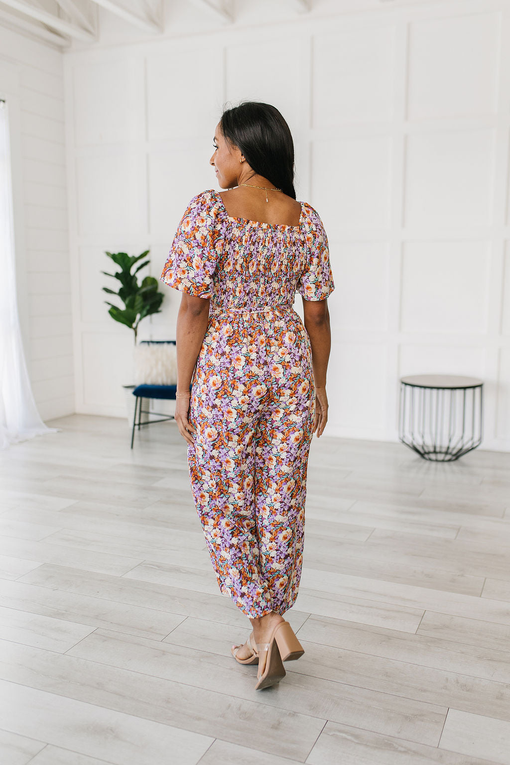 backview of Woman wearing a floral jumpsuit standing in a minimalistic room with white walls and light wood flooring.
