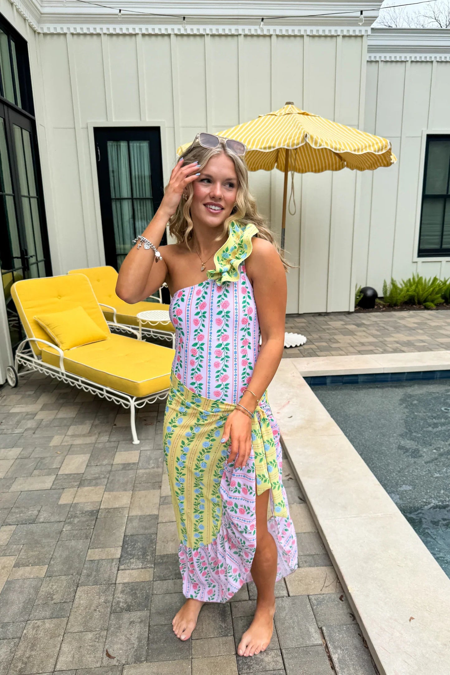 Woman in a colorful swimsuit dress standing by a poolside with a yellow umbrella and lounge chair.
