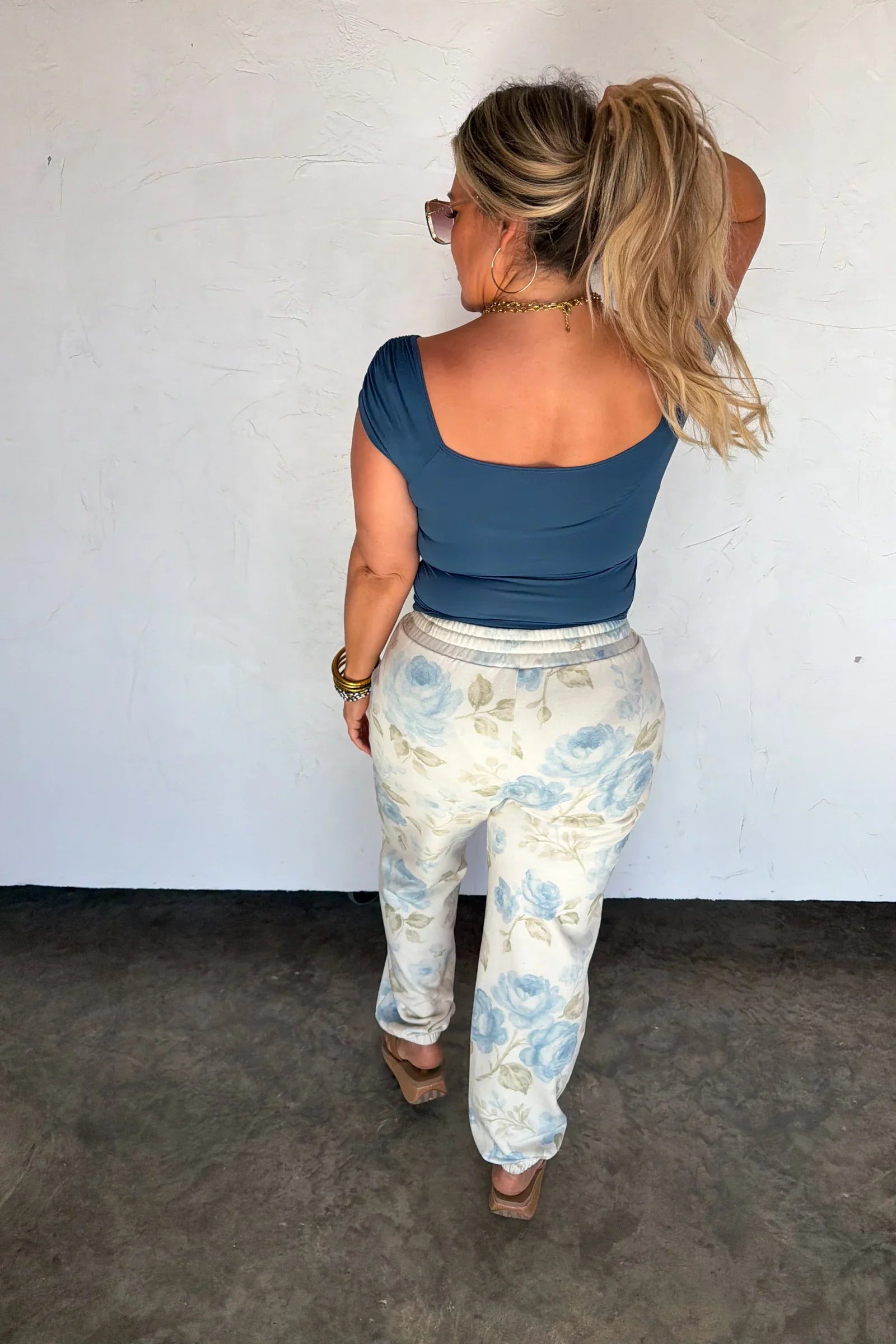 Woman wearing a blue top and floral pants against a white wall.