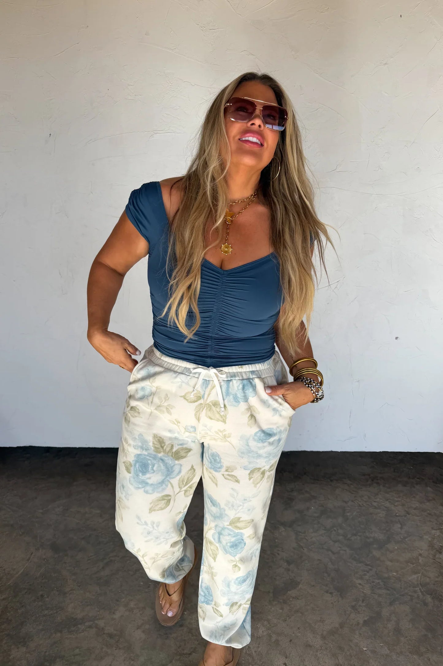 Woman wearing a blue top and bllue toile pants against a white wall.