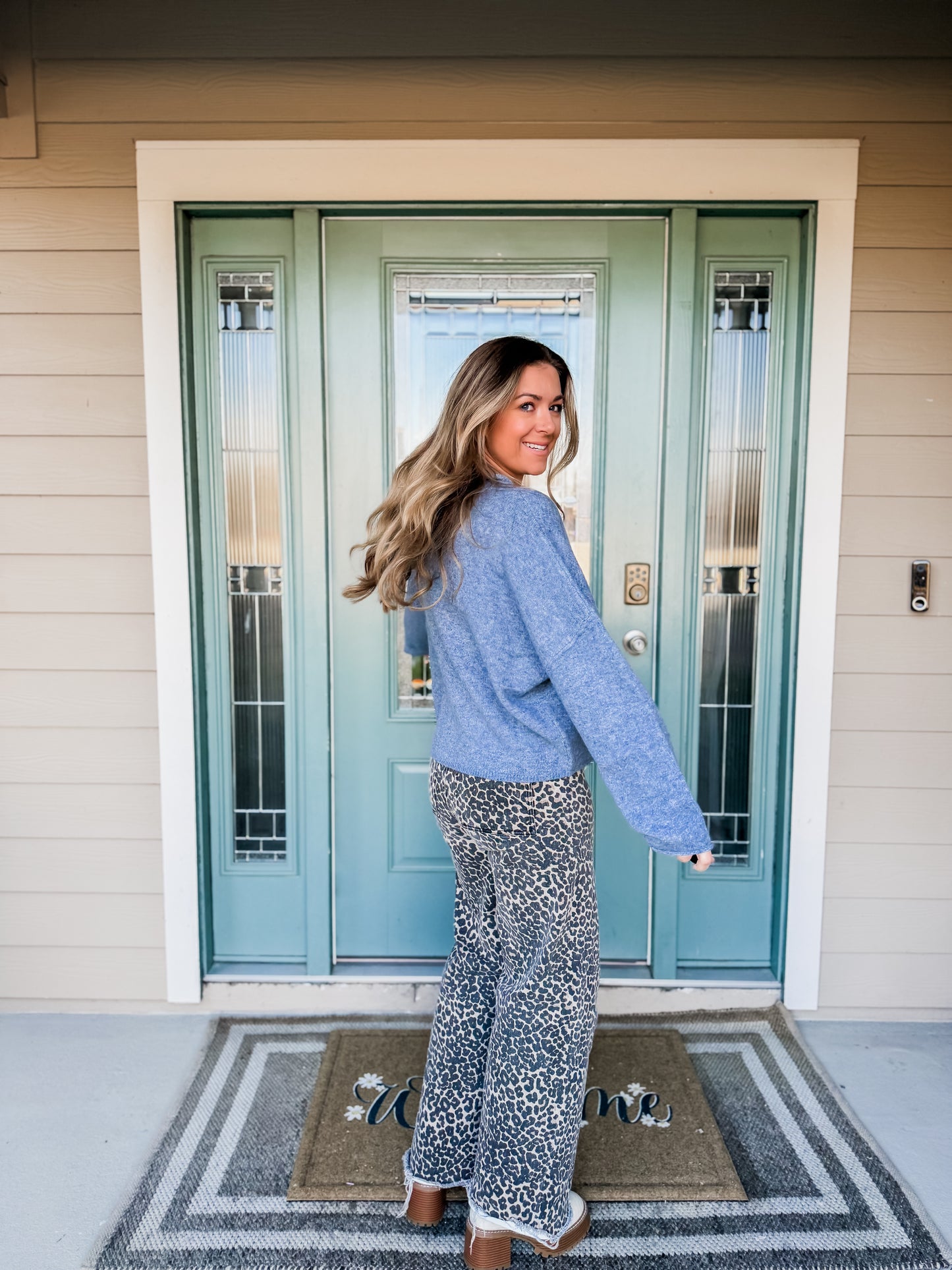 Woman standing on a porch with a light blue front door. back view of sweater called cozy up everyday cardigan in dusty blue. 