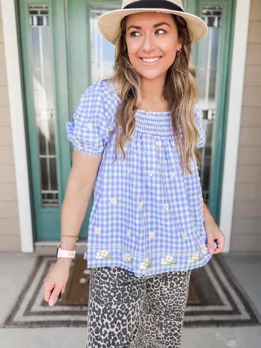 Woman wearing a blue checkered top with floral embroidery and leopard print pants, standing in front of a green door.