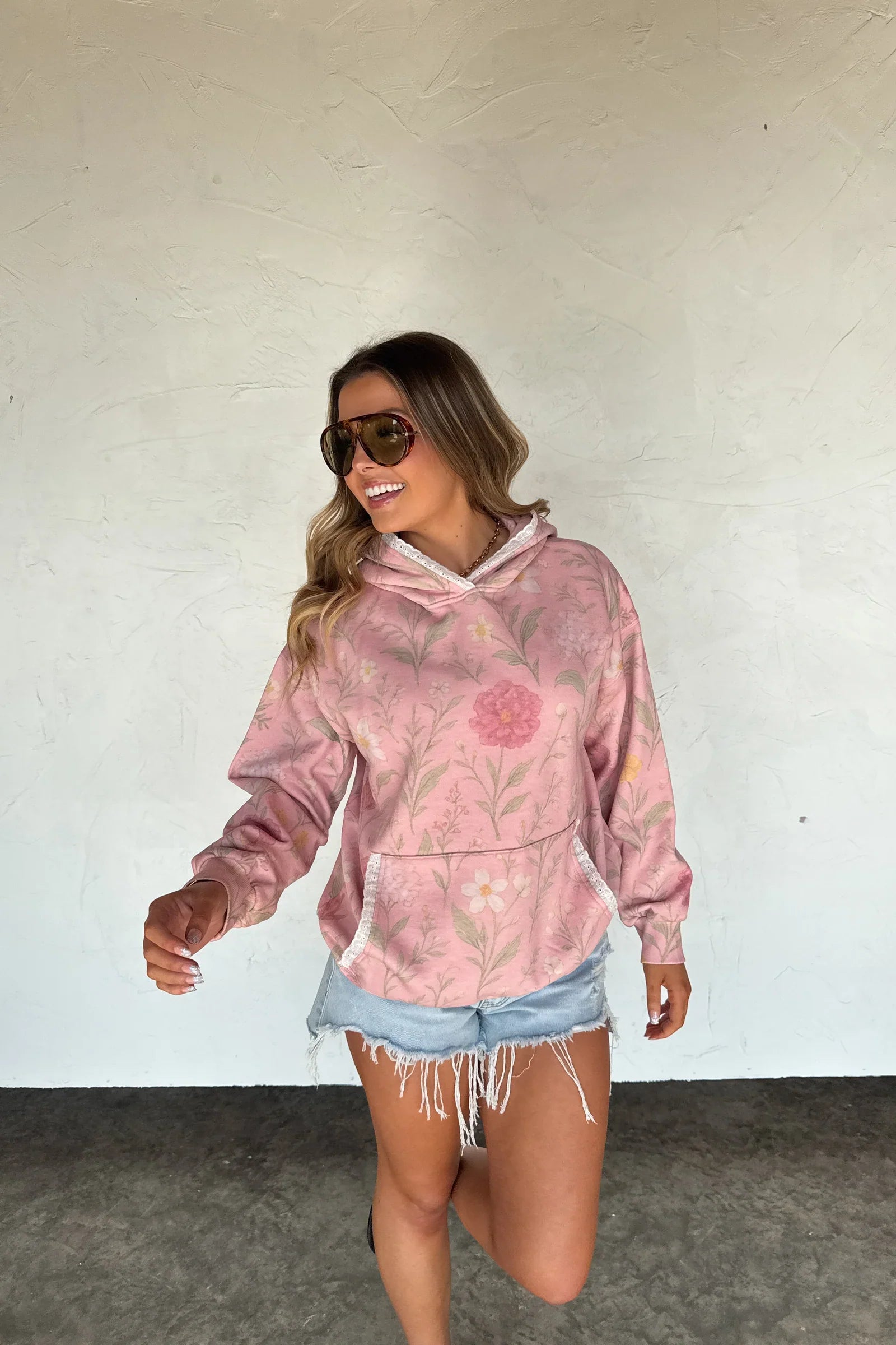 Woman wearing a pink floral hoodie and denim shorts against a white wall.