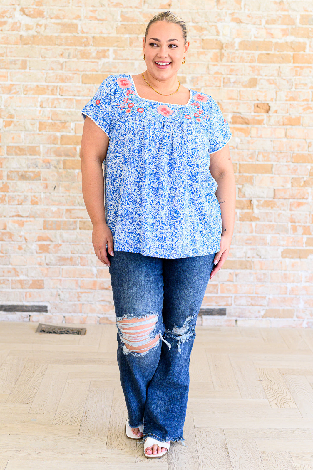 Woman wearing a blue floral top and ripped jeans standing against a brick wall.