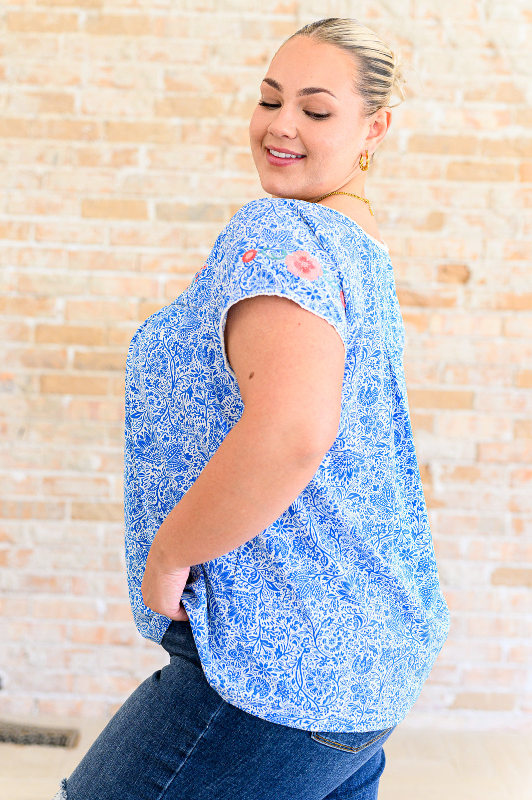Woman wearing a blue floral blouse against a beige brick wall.
