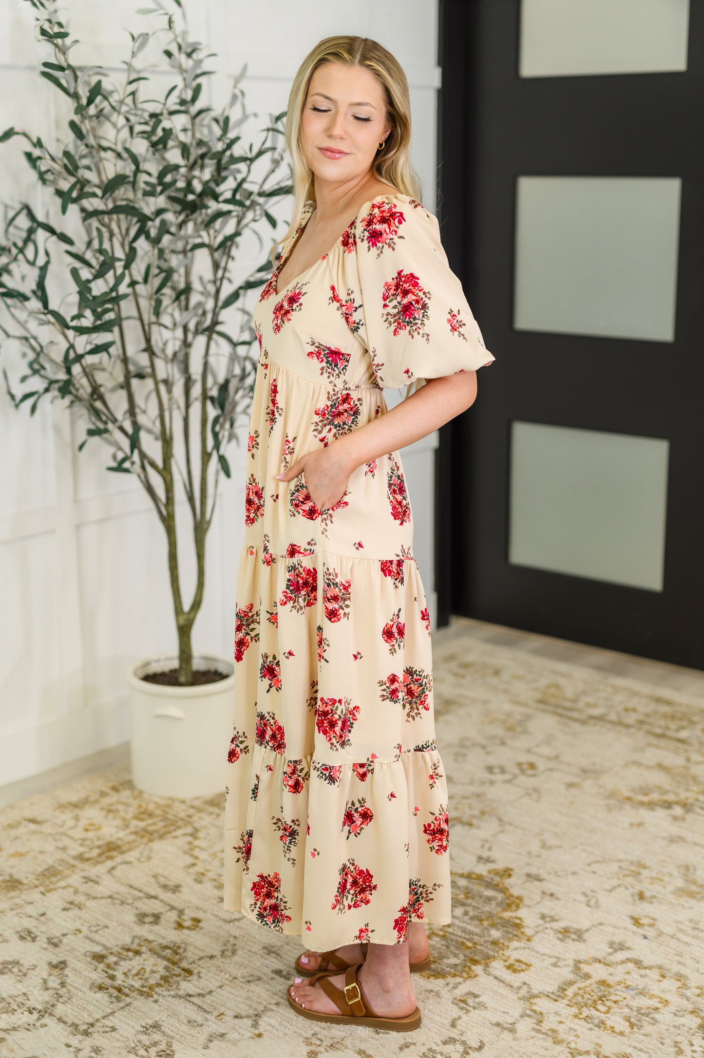 Woman wearing a floral dress standing in a room with a plant and decorative wall.