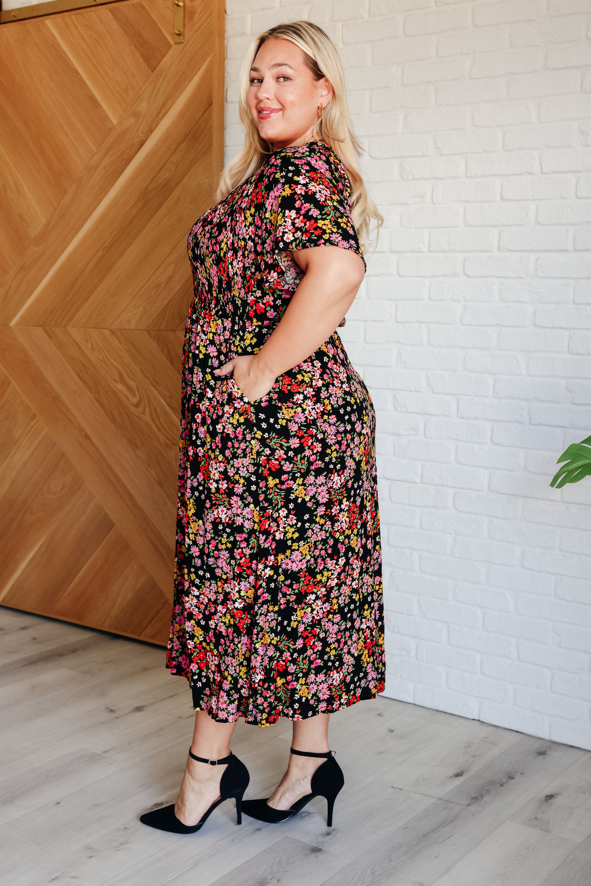 Woman wearing a floral dress standing against a wooden and white brick wall.